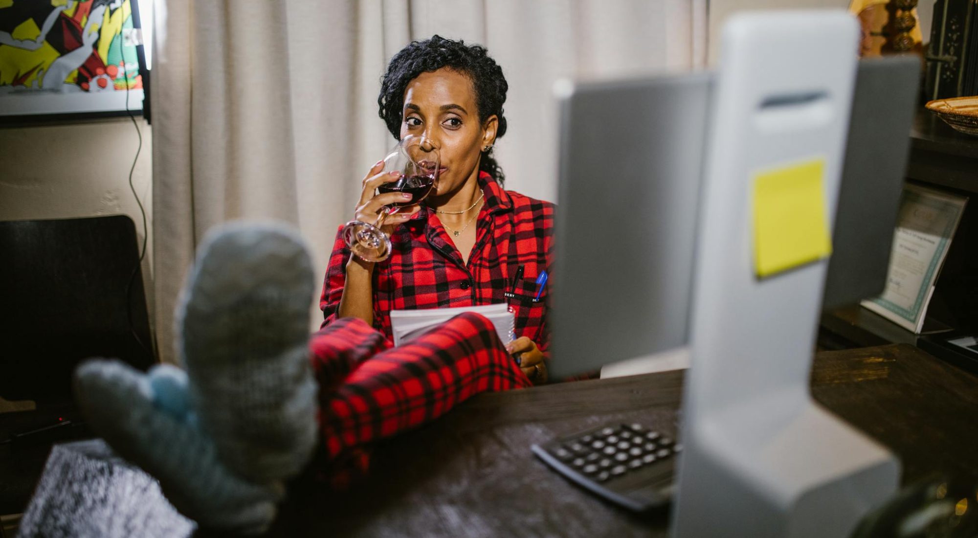 Woman in red pajamas relaxing with wine at a home office desk with feet up.